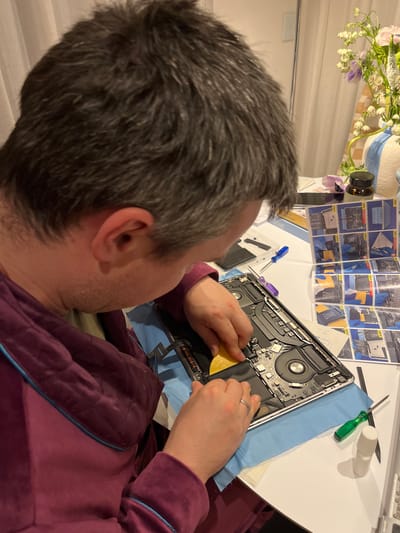 A man with graying hair is focused on repairing a laptop, open and disassembled, on a white table. He is using a yellow tool to work on the laptop's components, with a blue cloth underneath for protection. An instruction manual illustrating the repair steps is visible above the laptop. Various tools, including screwdrivers and a white container, are scattered on the table alongside the laptop. The man is wearing a maroon quilted robe. A vase of flowers sits on the left side of the table. There is no visual information to ascertain the city, Gangloff, Germany.