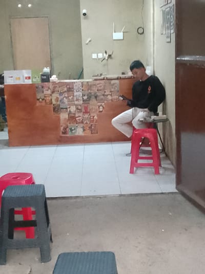 A young man wearing a black long-sleeved shirt and white pants is seated on a red plastic stool, holding a mobile device. He is positioned in front of a counter display adorned with various posters. The counter itself has a wooden surface and is lined with small boxes and bottles. The flooring is tiled. To the left of the man, a red plastic stool is visible, and to his right, a small table with items on it. In the background, a closed door and a wall with a surveillance camera are present. The scene appears to be indoors, possibly in a shop or cafe, within Cisarua, Indonesia.