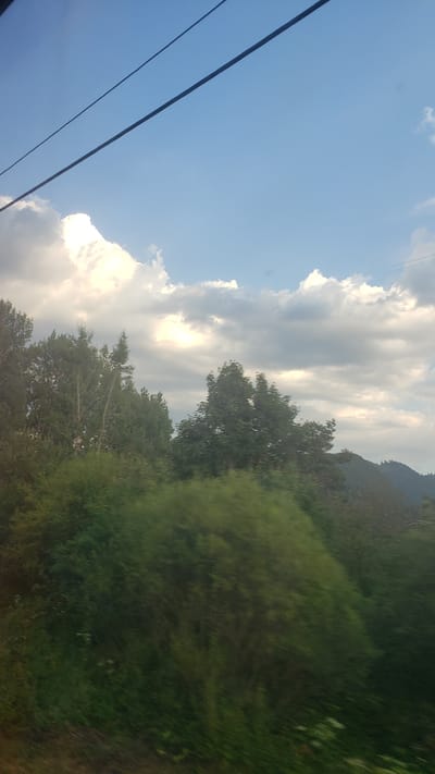 The image depicts a view from a moving vehicle, likely a train or car, looking out at a landscape. Two dark, parallel lines, possibly power lines or overhead cables, stretch across the upper portion of the frame against a bright blue sky dotted with white and gray clouds. Below the sky, a dense green treeline dominates the mid-ground and foreground, with some trees appearing taller and more defined than others. In the distance, rolling hills covered in trees recede into the background. The motion blur suggests the image was captured while in transit. The overall scene is one of a natural landscape viewed from a moving perspective. The location is identified as Hybe, Slovakia.