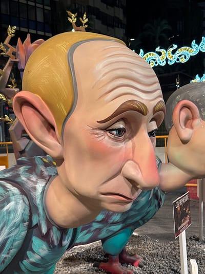 This image captures a close-up view of a large, artistic sculpture, likely part of a "Fallas" monument, in Alicante, Spain. The sculpture depicts a caricatured male figure with exaggerated features: a large, bulbous nose, prominent ears, and a furrowed brow with painted wrinkles. The figure's skin is rendered in a light, peachy tone with rouged cheeks. The eyes are blue with dark eyelashes and eyebrows. The figure wears a garment painted with a repeating pattern of teal and dark blue feathers. Behind this figure, another similar sculpture of a person's head is partially visible. The background shows a dark, urban environment at night, with building lights and decorative neon lights. A small sign in front of the sculpture reads "LA IMPERFECTA" with additional text below. The ground is covered with gravel.