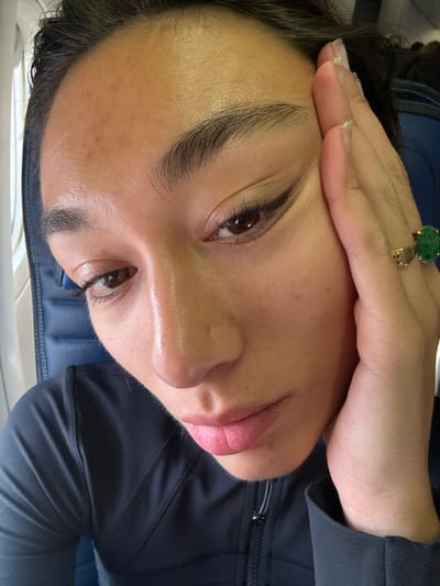 A close-up image depicts a person, appearing to be female, inside an aircraft cabin. The subject's face is prominent, featuring brown eyes with winged eyeliner, full eyebrows, and light pink lips. Her complexion shows some freckles or minor hyperpigmentation on the forehead. Her dark hair is pulled back. The subject's right hand, adorned with a gold-toned ring featuring a large, oval green gemstone on the ring finger, rests against the right side of her face near the temple. She is wearing a dark, long-sleeved athletic-style top with a zippered collar. In the background, elements of an airplane interior are visible, including a blue airline seat with textured upholstery and a window frame on the left. The specific location within Tremblay-en-France, France, is not discernible from the visual information provided in the image.