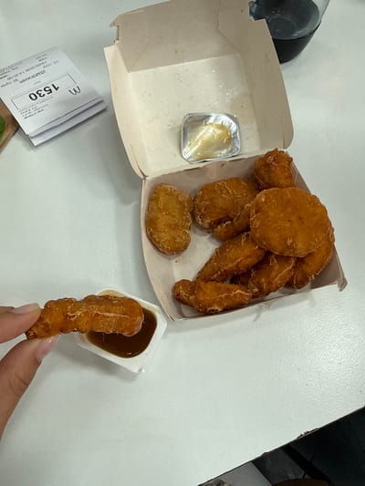 The image shows a McDonald's meal, likely chicken nuggets, on a white table. The food is in a partially open McDonald's container, with a small container of sauce inside. One nugget is being held by a person's hand and dipped into another sauce container. A McDonald's receipt with the number 1530 is visible, identifying the location as "Aice Mall - Kumat Hkanisu," Ariel Ltd., 8 Kampen, Eilat, Israel. The receipt indicates the date as 19/05/2026 at 14:35:06.