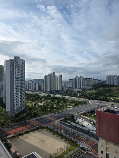 This is a high-angle, panoramic, exterior photograph of a residential area under construction and development in Daejeon, South Korea. The scene depicts a collection of modern, high-rise apartment buildings, some of which are completed and occupied, while others are still in various stages of construction. Cranes are visible amidst the new buildings, indicating ongoing construction activity.
In the foreground, a large, partially completed building is covered in reddish-brown netting, suggesting active construction. Adjacent to this, a sandy area with some sports equipment, possibly a makeshift soccer field, is visible, with a small shaded structure to its left. Several roads and intersections are present, with cars driving on them, indicating a functional transportation network. Pedestrian crossings with yellow and white markings are clearly visible. Green spaces, including trees and grassy areas, are interspersed throughout the urban landscape.
In the mid-ground, a multi-lane highway overpass crosses the scene, with more apartment buildings and infrastructure stretching into the distance. The sky is partly cloudy, with patches of blue sky visible. In the far background, rolling hills and mountains are discernible. The buildings are characterized by their rectangular shapes and numerous windows, typical of modern residential architecture. The overall impression is one of a rapidly developing urban area.