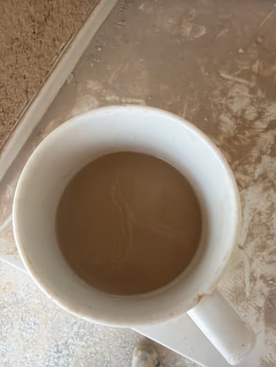 A close-up, overhead shot shows a white mug filled with a light brown liquid, likely coffee or tea, resting on a saucer. The mug has a subtle swirl pattern visible on the surface of the liquid. The mug and saucer are placed on a textured surface with a mottled brown and beige pattern, possibly a countertop or table. The background shows a continuation of this textured surface and a distinct edge with a rough, brown material, suggesting it might be near a window sill or a different material transition. There are no people visible in the frame, and the setting appears to be indoors, on a flat surface. The image does not provide any specific visual cues to pinpoint its location in Příbram, Czechia.