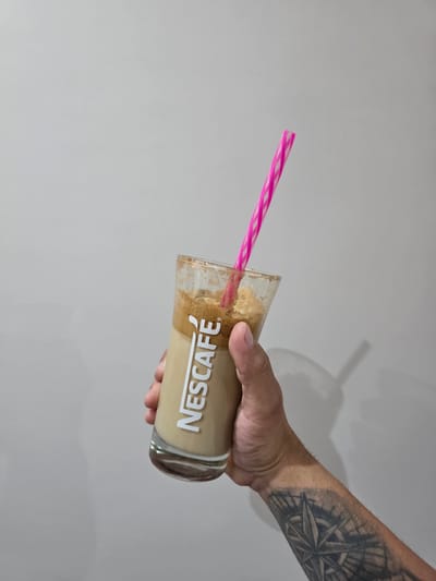 In the city of Vidin, Bulgaria, a close-up shot captures a hand holding a glass of a light brown beverage, possibly coffee, with a pink and white striped straw. The glass features a white "NESCAFÉ" logo. The beverage appears to have a creamy, foamy layer on top. The hand is tattooed with a compass rose design. The background is a plain, light gray wall, casting a faint shadow of the glass and straw.