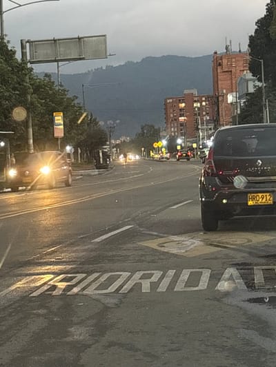 This image depicts a multi-lane urban road in Bogota, Colombia, at dusk or early evening under a heavily overcast sky. The asphalt road surface is wet. In the immediate foreground, the word "PRIORIDAD" (Spanish for "PRIORITY") is painted in white capital letters across the road, accompanied by a yellow square with a circular symbol.
On the right side of the road, the rear of a black Renault Sandero vehicle is stationary, displaying a Colombian license plate "HRD-912" with green reflective dots on its bumper. Further along this side, a multi-story red-brick building with several illuminated windows is visible. The distant background is dominated by dark, tree-covered mountains, characteristic of Bogota's Cerros Orientales.
On the left side of the road, a dark-colored sedan with brightly illuminated headlights approaches the camera in the opposing lane. Adjacent to this lane, a bus stop structure with a yellow sign (partially displaying "Vial") stands amidst green trees.
In the mid-ground, centered between the traffic lanes, a light-colored utility vehicle, possibly a small truck or SUV, is overturned on its passenger side, facing away from the camera. A garbage bin or similar container is located near the overturned vehicle. Further down the road, numerous vehicles with activated headlights and taillights are visible, indicating ongoing traffic. A solitary figure on a bicycle is faintly discernible in the distant lane, moving away. No emergency personnel or individuals are seen directly interacting with the overturned vehicle.