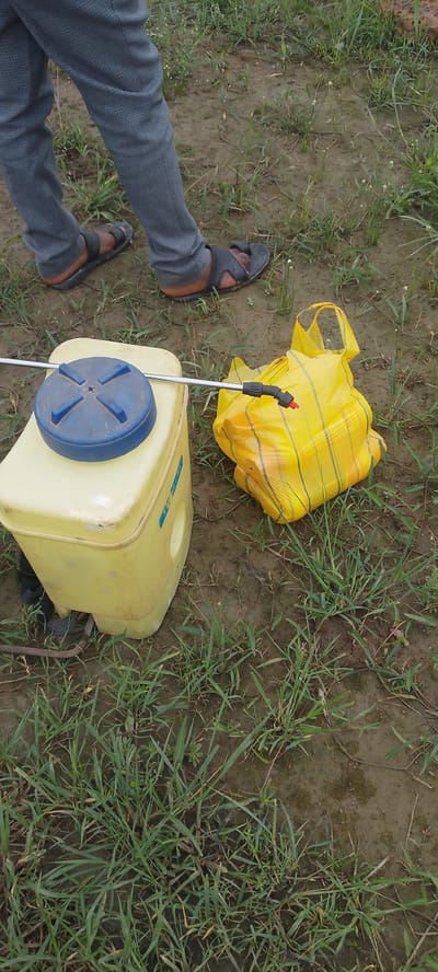 The image depicts a close-up view of a person's legs, clad in grey trousers and black sandals, standing on muddy ground with sparse green grass. To the right of the legs is a yellow plastic sprayer tank with a blue lid and a metal wand attached to it. Next to the sprayer, a crinkled yellow plastic bag with dark stripes is partially visible. The environment appears to be an outdoor agricultural setting, possibly a field, in Angwan Hausawa, Nigeria. The lighting suggests it is daytime.