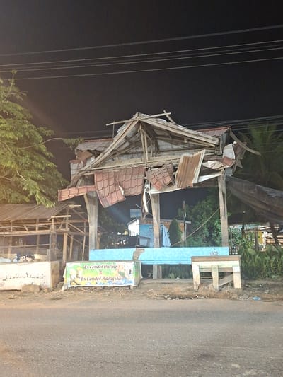 The image depicts a damaged outdoor structure in Keudeteupinpunti, Indonesia. The structure, possibly a food stall, has a partially collapsed roof made of corrugated metal and tiles. The wooden support beams are exposed and splintered. In front of the structure, a banner displays "Es Cendol Durian & Es Cendol Malaysia," indicating it was likely a place selling beverages. A low blue counter runs along the front, and a wooden stool or bench is positioned to the right. The ground in front of the stall is littered with debris. The background shows trees and other structures, partially obscured by darkness and the damaged stall. Power lines are visible overhead. The scene appears to be at night, with artificial lighting present. There are no people visible in the image.