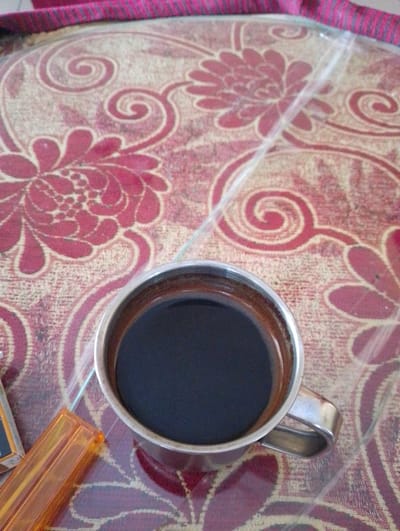 A close-up, overhead shot shows a metallic cup filled with dark liquid, presumably coffee, resting on a patterned surface. The surface features a repeating floral and swirling motif in shades of red and beige. To the left of the cup, partially visible, is an orange, transparent object, possibly a lighter. A thin, clear plastic sheet overlays part of the scene, creating a slight glare. The background suggests a tabletop or similar surface. The image was captured in Sukamanah, Indonesia.