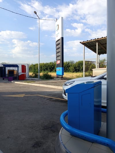 The image captures a view of a gas station in Tömönkü Norus, Kyrgyzstan. In the foreground, a white car is parked at a pump, partially obscured by a blue dispensing unit and a blue metal railing. Behind the car, a tall, illuminated sign displays fuel prices. To the left, a generator set is visible within a blue container, next to a red cabinet. The ground is asphalt with a yellow directional arrow. The background features a line of green trees and a field under a partly cloudy sky. The scene appears to be set during the daytime.