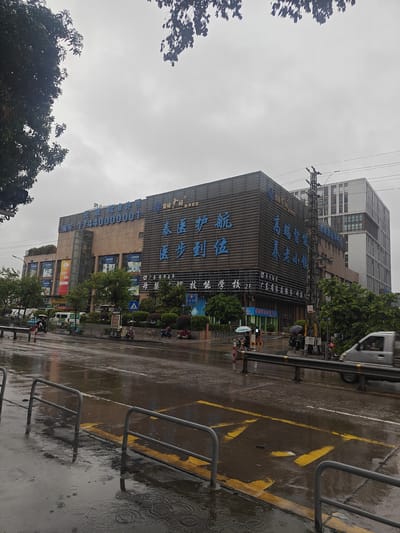 A low-angle, exterior, medium shot captures a multistory building with large blue Chinese characters on its facade, situated on a city street in Nanhai District, China. The overcast sky is visible above the building and trees. The street in front of the building is wet and reflects the surrounding lights, indicating recent rain or ongoing precipitation. Several parked motorcycles and scooters are visible on the sidewalk, along with a white van and a silver van driving on the wet road. Pedestrians are present, some holding umbrellas. Metal railings line the sidewalk, and yellow road markings are visible on the street. The building appears to be a commercial or residential complex, with various signs and advertisements displayed on its exterior. The scene conveys a somewhat damp and gloomy atmosphere due to the weather.