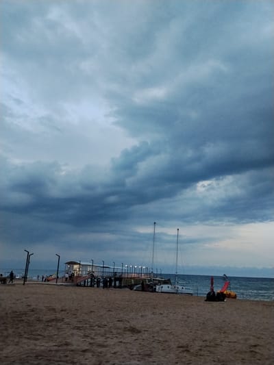 The image depicts a cloudy day at a beach, likely in the city of Bosteri, Kyrgyzstan, with the Issyk-Kul Lake in the background. A pier extends into the water, featuring a white building with a blue roof and illuminated lamps. Several people are visible on the pier and the sandy beach in the foreground. Two sailboats are moored near the shore, one of which has the Cyrillic lettering "МИФ" on its hull. Additionally, a red and yellow water slide is present on the beach. The sky is filled with heavy, dark clouds, suggesting an approaching storm or overcast weather.
