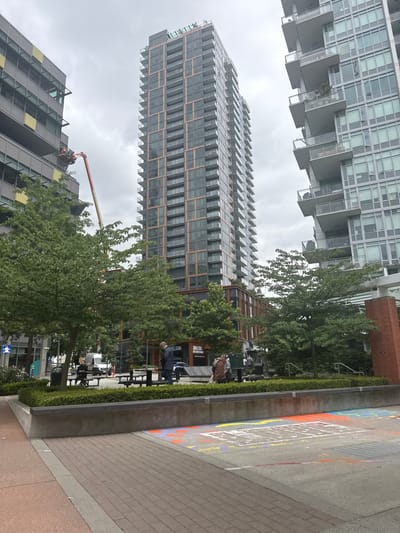 In New Westminster, Canada, a tall, slender skyscraper with a facade of glass and dark brown or bronze-colored vertical accents dominates the center of the image. To its right, another modern building of similar height features a repetitive pattern of balconies, most of which are enclosed with glass. To the left of the central skyscraper, a building with a grey facade and rectangular yellow accents is visible, with a red cherry picker extended towards it, reaching a mid-level section.
In the foreground, a paved area with brick-like patterns transitions into a concrete surface decorated with colorful chalk drawings, primarily in white, orange, and purple. A low, concrete planter with a dark-colored top border and a dense green hedge runs across the middle ground. Behind the hedge, several picnic tables are present, some occupied by people. A man in a dark jacket stands near one table, looking down at his phone. Another man, wearing a brown cloak-like garment, is seated at a table. A white van is parked in the background, and more trees and the facades of buildings are visible. The sky is overcast.