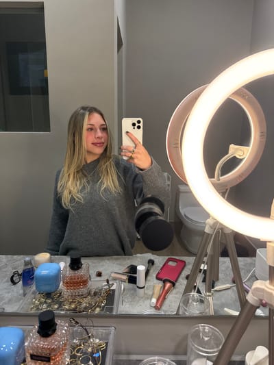 A female subject with long, highlighted blonde hair and light makeup is depicted taking a mirror selfie in a bathroom. The subject, wearing a grey crew-neck sweater, holds a white smartphone with a dark grey case and dark grey fingernail polish on her right hand, which also displays several gold-toned rings. Her gaze is directed slightly to her right, away from the phone's lens.
The reflection reveals a grey countertop beneath the mirror, cluttered with various personal items. These objects include two perfume bottles (one clear with "VALENTINO" visible, another light blue and opaque), multiple makeup products such as brushes, lip glosses, and compacts, and a clear tray filled with assorted gold-colored jewelry (necklaces, earrings, rings). A red smartphone case is also visible on the counter.
To the subject's right, a large, illuminated circular LED ring light on a light-colored tripod stand is prominently positioned. The background reflection, behind the subject's head, shows a plain grey wall. Further to the right in the reflection, partially visible behind the ring light, is a white toilet, confirming the bathroom setting. The specific location within the city of Monterrey, Mexico, cannot be determined from the provided visual data.