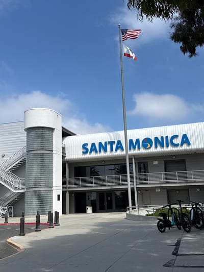 The image captures an outdoor scene in Santa Monica, California, under a clear blue sky with scattered clouds. The main subject is a modern building with a large blue sign that reads "SANTA MONICA". The sign features a colorful circular logo with shades of orange, yellow, and blue.

In front of the building, a flagpole stands tall, with two flags flying: the American flag and the California state flag. The flags are partially unfurled, suggesting a light breeze. To the right of the building, several bicycles are parked and locked to a rack. These appear to be shared electric bikes, as indicated by the "Veo" branding visible on one of them.

The building itself has a white, ribbed facade and features large windows and metal grating. The lower section of the building shows a modern architectural design with curved elements and a grid-like glass facade. The ground is paved with concrete and marked with yellow and red lines along the curb.

The overall impression is a sunny day in an urban environment, likely in the daytime given the bright lighting. The scene conveys a sense of normalcy and everyday activity, with the parked bikes suggesting transportation and public accessibility. No people are visible in the frame.