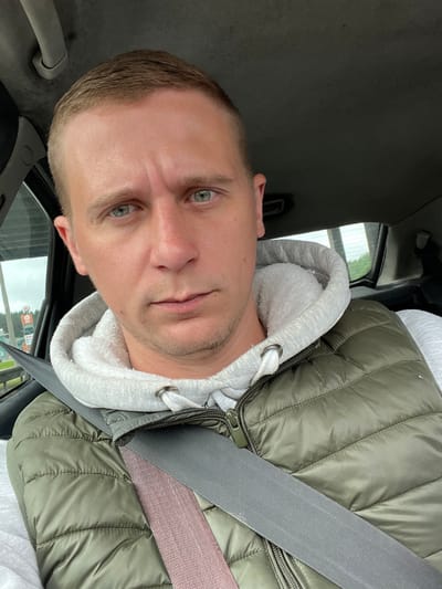 A man is sitting in the driver's seat of a car, looking directly at the camera. He has short, brown hair, light blue eyes, and a slight stubble on his face. He is wearing a light gray hooded sweatshirt with a white fuzzy lining and a dark green puffer vest over it. A grey seatbelt is fastened across his chest. The interior of the car is visible behind him, including part of the headliner and the passenger side window. Through the window, a blurry view of trees and a sign for a Burger King can be seen, suggesting the location may be near a roadside establishment in or around Бараўлянскі сельскі Савет, Belarus. The lighting indicates it is daytime.