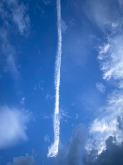 The image captures a vivid blue sky with scattered clouds. A prominent, bright white contrail streaks vertically through the center of the frame, appearing as if a jet plane has recently passed overhead. The contrail is thin and wispy at the top, gradually widening and becoming more textured towards the bottom. Surrounding the contrail are various cloud formations. To the left, there are softer, lighter clouds, some appearing almost transparent against the blue. To the right and towards the bottom of the image, darker, more ominous clouds are visible, hinting at changing weather conditions. The overall lighting suggests it is daytime, with the sun illuminating the contrail and the brighter clouds. There are no people, buildings, or specific landmarks visible to indicate a location within the city of Berkovitsa, Bulgaria.