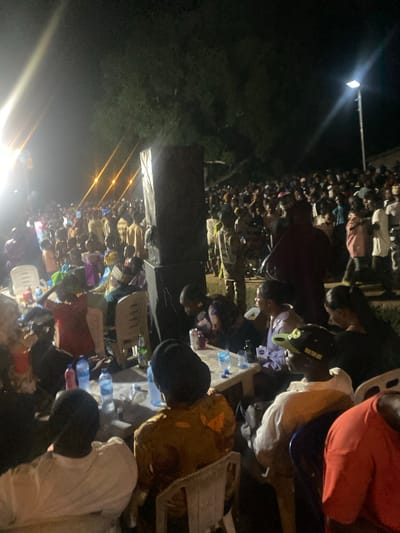 The image shows a large gathering of people outdoors at night. It is poorly lit, but a streetlight and some string lights are visible. Most people are sitting or kneeling, and there are many plastic water bottles scattered around. People are wearing different colors of shirts and garments. The location is identified as Epe, Nigeria.