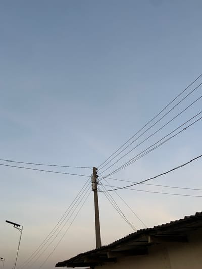 In Barikin Jirga Summa, Nigeria, the image captures a utility pole standing tall against a pale blue sky. Multiple dark, thin wires crisscross the frame, emanating from the pole and stretching towards the right edge of the image. A section of a corrugated metal roof, belonging to an unseen structure, occupies the lower right portion of the scene. To the left, a modern street lamp with a long, slender pole and a dark, rectangular fixture at its apex is visible. The sky is clear and vast, indicating an open environment, likely on the outskirts of a residential or commercial area within Barikin Jirga Summa. There are no people or animals present in the frame.