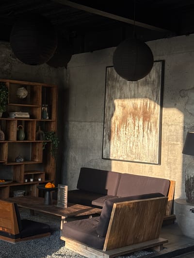The image presents an overhead perspective of an interior space, potentially a lounge or seating area. The location is suggested to be Pererenan, Indonesia, though no explicit landmarks confirm this.

Dominating the view are furniture arrangements including a cushioned sofa with a wooden table, a wooden shelving unit, and two hanging spherical lanterns. The shelf displays books, decorative items, and plants.
The light source appears strong, casting shadows and highlighting textures on the walls and furniture.
The scene is static; there are no people or visible actions.