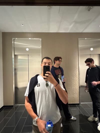 The image depicts three males in a modern interior hallway, likely a hotel or residential building lobby or common area, located in the city of Boston, United States. The scene is captured as a mirror selfie, with the primary subject holding a dark-colored smartphone to take the photograph.
The foreground subject is a male with short, dark hair, wearing a white t-shirt with a circular badge on the left sleeve (identifiable as Stone Island), a dark sweater draped over his shoulders, and sunglasses hanging from his collar. A silver wristwatch is visible on his left wrist, and he holds a blue, rectangular carton or bottle in his right hand.
Reflected in the mirror to the left of the foreground subject, slightly behind him, is a second male with dark, curly hair and eyeglasses, wearing a dark t-shirt with a graphic design and holding a clear plastic water bottle. To the right of the foreground subject, also reflected in the mirror and positioned further back, is a third male with dark, curly hair and sunglasses, dressed in a dark t-shirt and dark pants, holding a clear bottle.
The environment includes two stainless steel elevator doors on a light beige wall. The floor is composed of dark, rectangular tiles. Overhead, recessed lighting is visible in the white ceiling. The subjects appear to be waiting or passing through the area.