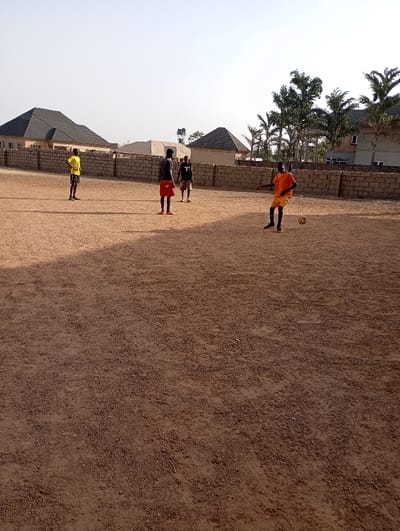 Four people are playing soccer on a dirt field in front of a low brick wall. Three are standing near the wall, while one in an orange shirt is kicking the ball. Behind the wall are houses with dark roofs and palm trees. The field takes up most of the frame and has a dry, cracked surface. It is daytime and the sun is shining.