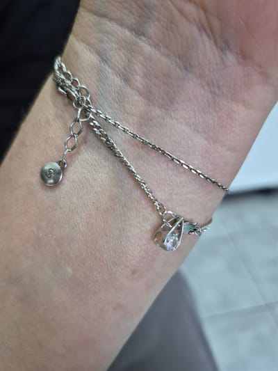 A close-up, overhead shot shows a delicate silver bracelet on a person's wrist and forearm. The bracelet consists of two thin chains: one with a small, round charm engraved with a stylized letter 'S', and another adorned with a teardrop-shaped pendant holding a clear, faceted gemstone. The jewelry rests on lightly tanned skin with visible veins and subtle creases. The background is blurred, revealing a dark garment on the left and a light, textured surface on the right, possibly fabric or a wall. There is no identifiable information about the location of Nyagan, Russia in the image.