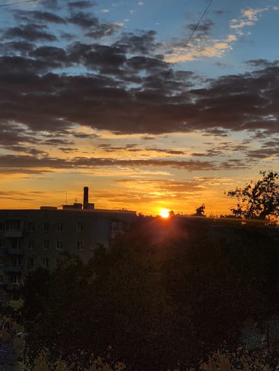 The image shows a sunset over the city of Murom, Russia. The sky is filled with a mix of dark clouds and bright orange and yellow hues, indicative of the sun setting. In the foreground, there is a silhouetted apartment building with visible windows and a chimney stack rising from one of its sections. Trees with dark foliage are obscuring the lower part of the building and the ground. A single tree with a fuller canopy is visible on the right side of the image, also silhouetted against the sunset. Overhead, thin wires are stretched across the frame, suggesting the presence of electrical or communication lines. The overall scene depicts an urban environment at dusk, with natural light phenomena creating a dramatic backdrop.