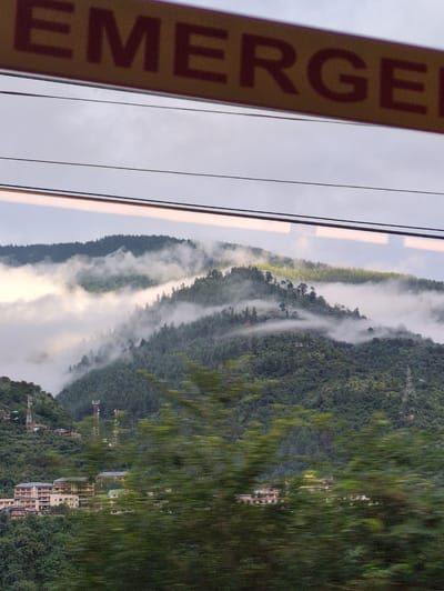 An eye-level shot shows a mountainous landscape with a band of yellow caution tape across the top, reading "EMERGENCY" in dark red letters. The mountains are covered in dense green trees and patches of white mist are swirling around them, giving the impression of a cloudy day or a foggy morning. Several buildings are visible on the lower slopes of the mountains, including a cluster of multi-story structures that appear to be hotels or apartment complexes. Communication towers are also present on the hillsides. The foreground is blurred, suggesting the photo was taken from a moving vehicle. The overall impression is of a scenic, slightly overcast day in a mountainous region. The location is identified as Chattan Seri, India.