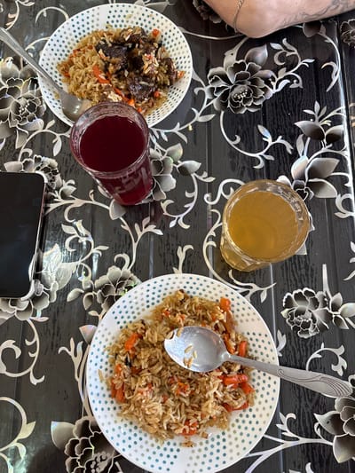 This image shows a table setting with two plates of rice pilaf, each with a spoon, along with two glasses of liquid, one clear and one red, and a smartphone. The pilaf on the left appears to contain meat, while the pilaf on the right appears to contain carrots. The table is black with a white floral pattern. The location is not evident from the image, and there is no indication it is in the city of Ust-Ilimsk, Russia. An arm is visible in the upper left corner of the photo.