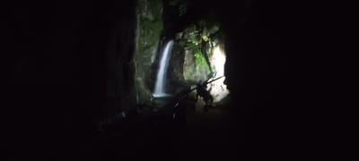 The image depicts a dimly lit scene within a cave or rocky overhang, featuring a waterfall cascading down a moss-covered rock face. The water is illuminated, appearing as a white stream against the darker surroundings. In the foreground, a wooden railing or barrier is visible, and a dark silhouette of a person, possibly standing or walking, can be discerned near the railing, facing the waterfall. The environment appears natural, with lush greenery clinging to the rocks. This scene is located at the Stobski Monastery Waterfalls, near the town of Kostenets, Bulgaria.