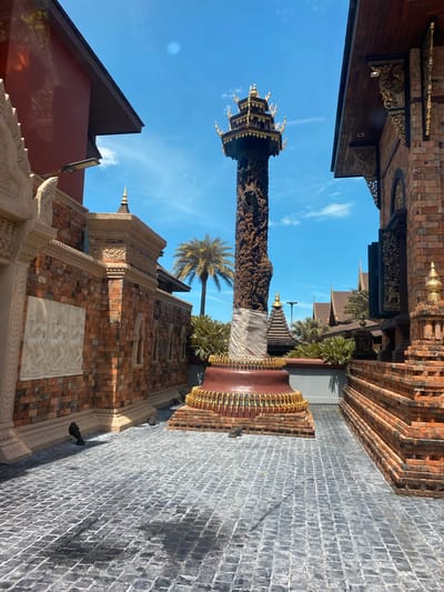 The image depicts a courtyard area with intricate architecture, likely a temple or cultural site, located in Baan Lamai, Thailand. A tall, intricately carved wooden pillar stands prominently in the center, topped with a multi-tiered golden structure. The pillar is partially wrapped in white cloth near its base, which is adorned with a reddish-brown pedestal featuring golden decorative elements and a brick base.

Surrounding the central pillar are buildings constructed with reddish-brown bricks, exhibiting detailed carvings and ornamental features. To the left, a building with a dark reddish-brown exterior wall and a dark brown roofline is visible, with a modern-looking light fixture attached to its facade. Adjacent to this is a section of brick wall with decorative light-colored relief carvings depicting figures in a dance-like pose.

To the right, a stepped brick structure with a layered appearance leads up towards a building with dark wooden shutters and intricate golden carvings. The ground is paved with dark grey cobblestones arranged in a diagonal pattern, with shadows cast across it, suggesting a sunny day. In the background, palm trees and other lush greenery are visible, along with the tiled roofs of other structures, indicating a tropical environment. The sky is a clear, bright blue with a few wispy white clouds. The overall scene conveys a sense of cultural heritage and architectural splendor under a bright, tropical sky.