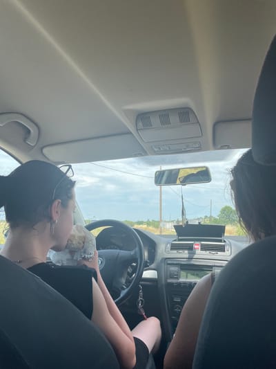 The image depicts the interior of a car, viewed from the back passenger seat. A young woman with dark hair tied up is driving, wearing a black top and glasses perched on her head. She is holding a wrapped sandwich in her hands and appears to be taking a bite. A silver necklace is visible around her neck, and she has a long, thin earring in her left ear. The car's dashboard is visible, including the steering wheel, radio, and air vents. Outside the car, the sky is overcast with some blue patches, and rural landscape with trees and power lines can be seen. This scene likely takes place in Myrodato, Greece, given the context provided.