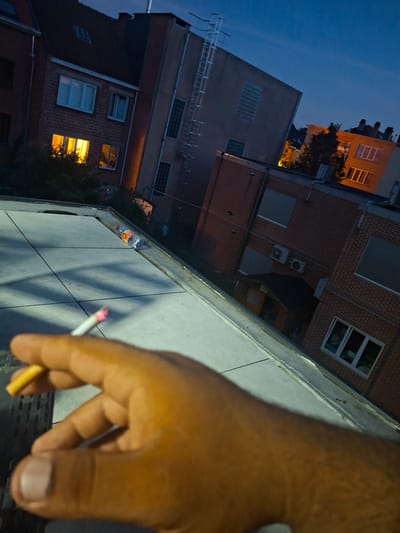 The image captures a person's hand holding a lit cigarette, with the tip glowing red, in the foreground. The hand is large and brown-skinned. The scene appears to be outdoors, possibly on a rooftop or balcony, as evidenced by the concrete wall with visible tile lines in the mid-ground and a railing-like structure in the background.
Behind the hand and wall, the view extends to several buildings. These buildings are constructed with brick and have various windows, some illuminated from within, suggesting it is nighttime or dusk. One building has a prominent orange glow from its windows, indicating interior lights. Another building in the background has a ladder-like structure on its exterior. The sky above is a deep blue, characteristic of twilight or a clear night.
The overall mood is somewhat relaxed and possibly contemplative, given the presence of a person smoking. The lighting suggests a time around dusk or early evening. There is no visible text in the image. The location context of Evere, Belgium, does not provide specific visual cues within the image itself, but places the scene within a European urban environment.