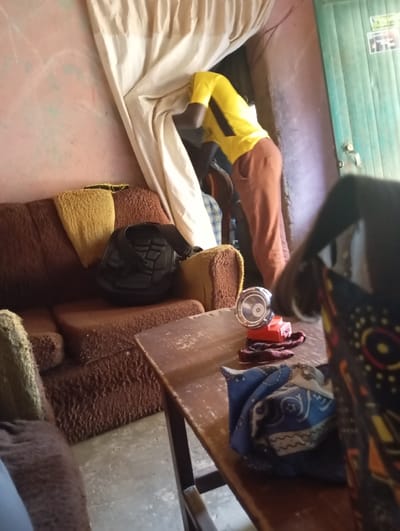 An indoor scene in Reifild-Ist, Nigeria, depicts a person in a yellow shirt with black accents and brown trousers bending forward behind an off-white draped fabric, their head obscured. To the left, a brown textured sofa with yellow fabric overlays holds a black object. In the mid-ground, a dark brown wooden table, exhibiting wear, features a silver flashlight with a red base, a small red fabric item, and a folded blue and white patterned cloth. The room's walls are a light reddish-pink with visible white markings, and the floor is light grey concrete. A teal door with a silver handle and stickers is visible in the background right.
