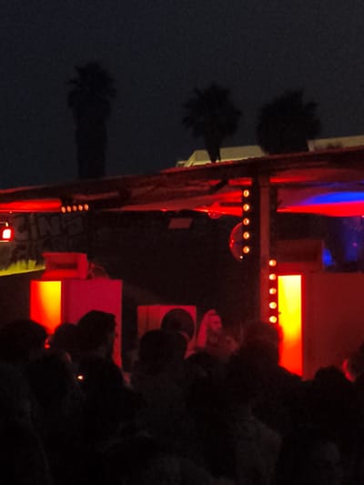 The image depicts a night scene at a venue, likely an outdoor event or concert, in Sant Adrià de Besòs, Spain. Silhouettes of palm trees are visible against a dark sky in the background. The foreground is dominated by a crowd of people, their forms largely obscured by darkness and the red and blue lighting. A stage or structure is lit with vibrant red and yellow lights, featuring what appear to be illuminated boxes or platforms. One of these platforms displays the word "CINA" in large, stylized lettering, suggesting a possible connection to cinema or a specific event name. The overall atmosphere is one of a lively, possibly music-focused, gathering, illuminated by artificial lighting that casts a dramatic glow on the scene.