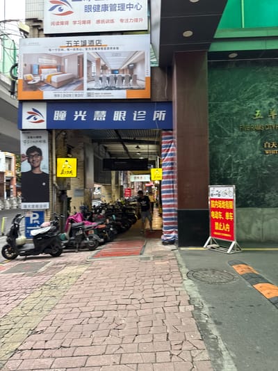 The image shows a street scene in Yuexiu District, China, featuring a row of parked motorcycles and scooters. In the background, several signs are visible, including one for "五羊城酒店" (Five Rams City Hotel) and another for an "eye clinic." A red and white striped banner hangs on a building. The ground is paved with red bricks, and a yellow line marks a path. A blue parking sign with a white 'P' is also visible. The overall environment appears to be an urban commercial area.