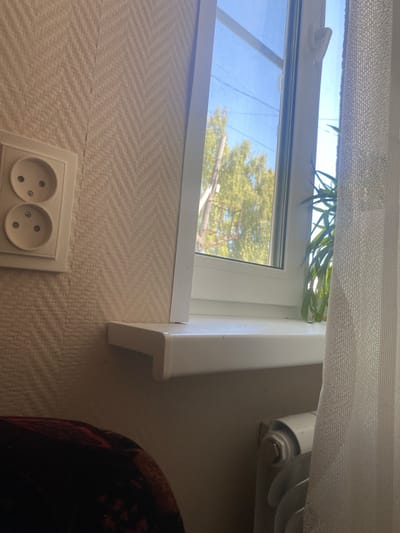 This image shows an interior view looking out a window. The window is partially covered by a sheer white curtain. Through the window, green trees and a blue sky are visible, suggesting it is daytime. A white windowsill runs along the bottom of the window. To the right of the window, a white radiator with a curved design is partially visible. Below the window, on a wall with textured wallpaper, is a double electrical outlet with two sockets. To the far right of the image, a portion of a dark, patterned blanket or piece of furniture is visible. The scene appears to be in a domestic setting, likely a room in a house or apartment in Afanasyevo, Russia. There is no discernible activity taking place, and the overall mood is neutral and ordinary. No text is visible in the image.