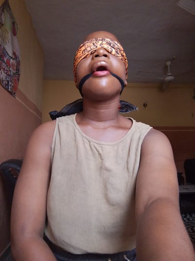 A medium shot shows a person with dark skin, wearing a beige sleeveless top, blindfolded with an orange and brown patterned cloth and a black strap around their mouth. The person's head is shaved. They are indoors, likely in a room in a house, judging by the wall color, a hanging decorative item on the wall to the upper left, and a ceiling fan in the background. The location is Umuahia, Nigeria, though no specific details in the photo confirm this location. There are other objects visible in the background. The person is looking upwards with their mouth slightly open. The person's skin appears moist or sweaty.