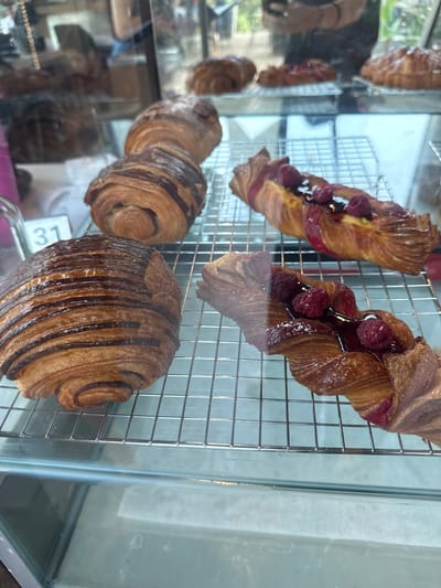 A display case holds various pastries. At the top are two twists with raspberries. Below them are three chocolate croissants. The location is San Pedro Garza García, Mexico.