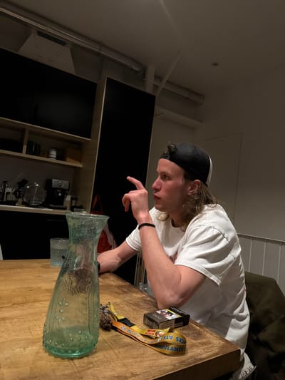 The image depicts an indoor scene, likely a residential kitchen or dining area, located in Saint-Denis, France. A single male individual is seated at a light-colored wooden table, positioned to the right of the frame. He has medium-length, wavy light-colored hair, and wears a black and white trucker-style baseball cap reversed. He is dressed in a white short-sleeved t-shirt. His left arm is bent, with his index finger extended upwards and forward, and his mouth is slightly open, indicating he is speaking or making a point. A black wristband is on his left wrist.

On the table in front of him, from left to right, are: a light greenish-blue glass carafe with embossed patterns, a partially visible clear glass, a yellow lanyard displaying text including "ATS AMBULANCE" and numbers, a keyring with a dark, grenade-shaped charm, and a red and white pack of Marlboro cigarettes.

The background reveals a modern interior. To the left, a wall-mounted flat-screen television is positioned above a light-toned wooden shelving unit containing various kitchen appliances, including a kettle and a coffee machine. A large, dark, vertical panel (possibly a cabinet door or wall feature) is to the right of the shelving unit. The ceiling features exposed white pipes and a structural beam with visible rivets. In the far right background, a white railing or banister is visible. The scene is illuminated by ambient overhead lighting.