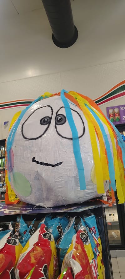 The image shows a piñata with a crude cartoon face and colorful crepe paper fringe on display for sale. The piñata is located in a 7-Eleven convenience store in San Miguel Acapantzingo, Mexico. It is situated next to bags of Sabritas brand potato chips. A ventilation duct is visible on the ceiling.