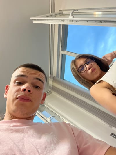 An upward-angle selfie-style photograph depicts two young individuals indoors, in Karavelovo, Bulgaria. In the left foreground, a male with a short buzz cut and light facial hair on his chin gazes directly at the camera, his lips slightly pursed. He wears a light pink t-shirt with a white palm tree graphic on the chest. In the upper right background, a female with long brown hair and black-rimmed glasses also looks towards the camera, her lips forming a pout. She wears a white sleeveless top. Behind the subjects, a white-framed window is open, revealing a clear blue sky. Vertical blinds or pleated shades are visible on the right side of the window frame.