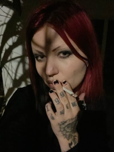 A close-up, chest-up portrait shot features a young adult female in Kunje, Montenegro. She has shoulder-length dark red hair, pale skin, and heavy dark eye makeup. A small silver ring is present in her left nostril. A distinct vertical shadow falls across the left side of her face. Her left hand is visible, holding a lit cigarette between the index and middle fingers, positioned near her mouth. Her fingernails are painted black, with some chipping evident. The fingers of her left hand display black script tattoos, and a larger black tattoo resembling a floral or mandala pattern covers the back of her hand. A black ring with an irregular shape is on her left ring finger. She wears a black long-sleeved garment. The background is dimly lit, with the shadows of bare tree branches cast onto a lighter surface on the left side of the frame, while the right side remains dark.