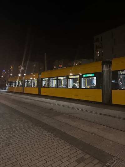 A yellow tram travels along a street at night. The tram is well-lit, and passengers can be seen through the windows. The surrounding buildings are visible in the background, with streetlights casting illumination on the scene. The ground is paved with cobblestones or bricks, and there is a set of tram tracks next to it. "7 Görlitz" is written on the front of the tram.