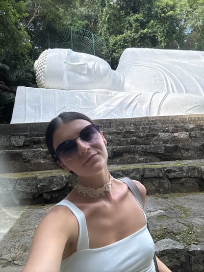A young woman with dark hair and sunglasses is taking a selfie in front of a large white reclining Buddha statue. She is wearing a white tank top and a lace choker necklace. The statue is on a stone platform, and stairs are visible in the foreground. The background is filled with lush green trees. The scene is likely located in the city of Thuận Nam, Vietnam, given the presence of a large Buddha statue.