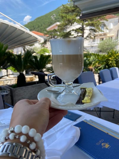 An outdoor cafe scene in Perast, Montenegro, features a hand holding a glass of latte with foamy milk. The latte is served on a white saucer with a spoon and two small, individually wrapped sweets – one dark brown and one yellow. In the background, a lush green mountain slopes upward, dotted with white buildings with red-tiled roofs. Palm trees and other greenery are visible in the middle ground, along with outdoor seating and tables draped with white tablecloths. The sky is a clear blue with a few white clouds. In the foreground, a blue menu with gold lettering that reads "CONTE" and some star symbols is placed on a white tablecloth.