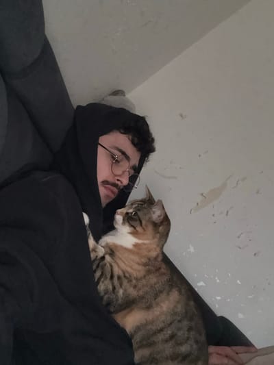 An indoor scene within a domestic residence in Rennes, France, depicts one adult male and one cat. The male, with dark curly hair, a mustache, and wearing round eyeglasses, is dressed in a black hooded sweatshirt with the hood up. He is reclined against a dark gray upholstered surface, his head slightly tilted downward, with his gaze directed towards the cat. The cat, a tabby with brown, black, orange, and white fur, is positioned against the man's left side. One of the cat's front paws rests on the man's sweatshirt. The cat's head is turned to its right, looking upwards and away from the man. The background consists of a light-colored wall with multiple irregular patches of peeling paint or surface texture.