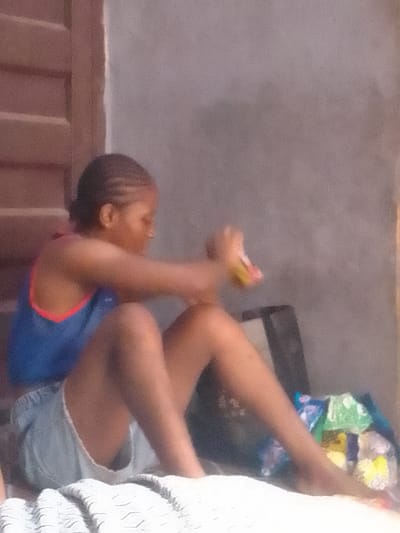 In Nkwelle Ezunaka, Nigeria, a young female is seated on a light-colored, patterned textile surface. She wears a blue tank top with an orange trim and light blue denim shorts. Her hair is braided and pulled back. Both hands are engaged with a small, multi-colored object, appearing to manipulate or open it. To the left, a wooden structure with horizontal slats is visible, likely a door or window shutter. To the right, a plain, light grey wall forms the background, along with a dark, indistinct object and a cluster of colorful plastic packages or bags. Her bare feet are visible on the textile surface.