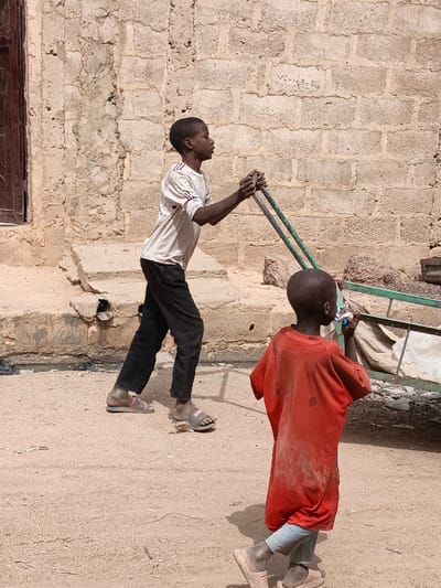 The image captures an outdoor scene in Zango, Nigeria, under bright daylight. Two males, appearing to be children, are present on an unpaved, light brown dirt ground.

The foremost individual, an adolescent male, wears a light-colored, short-sleeved t-shirt with dark stripes on the sleeves, and dark long pants. He has dark-toned skin and short hair. He is actively pushing or pulling a green metallic two-wheeled frame using both hands, positioned in front of him. His body is angled towards the right, indicating movement in that direction. His feet are clad in dark-toned slide sandals.

Behind him, a younger male child, also with dark-toned skin and short hair, walks in the same direction. He wears an oversized, dirty red short-sleeved t-shirt over light-colored long pants and light-toned slide sandals. He holds a small, multi-colored object near his mouth, looking slightly to his left.

The background consists primarily of a weathered, light brown concrete block wall. A dark brown wooden door or window frame is visible on the far left section of this wall. A raised concrete ledge or foundation, with a dark circular opening (possibly drainage) and dark fluid below it, runs along the base of the wall. Faint graffiti or markings, including "VCH 13 2B," are visible on the wall's surface. To the far right, partially obscured, is a portion of another metal frame structure, with some white, possibly fabric, debris underneath.